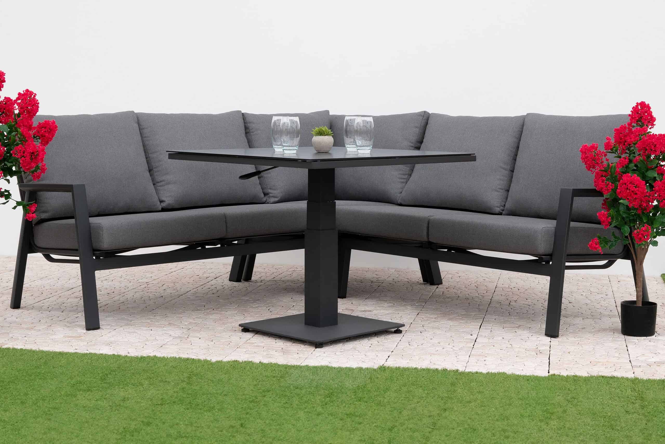 A photorealistic image of a sleek, modern grey aluminium garden dining set with a slatted tabletop and comfortable upholstered chairs. The set is positioned on a neatly paved patio in a medium-sized, aspirational UK garden during late autumn. Mature trees with vibrant red, orange, and golden yellow foliage are in the mid-ground, and borders feature late-flowering hydrangeas in muted pinks and purples alongside ornamental grasses. Soft, warm golden hour light filters through the trees, casting long shadows and a warm glow on the dining set and autumn leaves. The wide shot, slightly elevated composition places the dining set as the central focal point, slightly off-center according to the rule of thirds, with a beautifully blurred autumn garden background. The dominant color palette includes rich autumn tones of burnt orange, deep red, golden yellow, muted greens, and charcoal grey, with subtle hints of soft purple and pink.